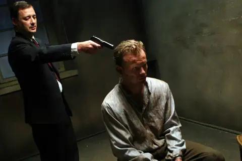 Scena z filmu "Generał Nil". Mężczyzna w garniturze celuje z pistoletu do siedzącego w brudnej koszuli więźnia.