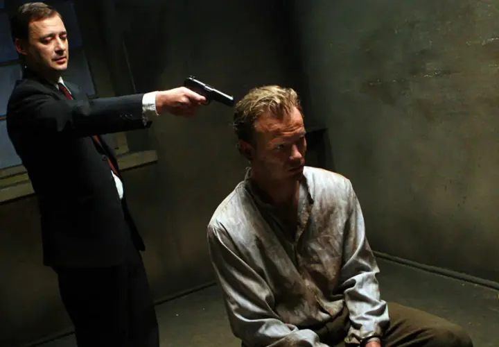 Scena z filmu "Generał Nil". Mężczyzna w garniturze celuje z pistoletu do siedzącego w brudnej koszuli więźnia.