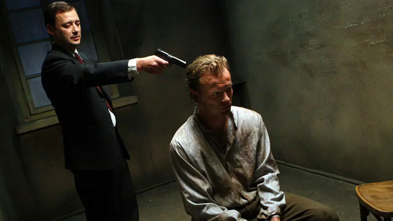 Scena z filmu "Generał Nil". Mężczyzna w garniturze celuje z pistoletu do siedzącego w brudnej koszuli więźnia.